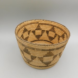 May include: A handwoven basket with a round shape and a wide opening. The basket is made of natural-colored fibers with a geometric pattern in shades of brown. The design features repeating diamond shapes around the sides.