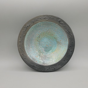 May include: A black ceramic bowl with a teal glaze and a black rim. The rim is decorated with a carved design.