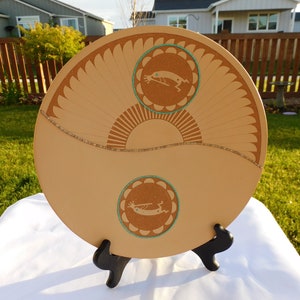 May include: A tan ceramic plate with two stylized figures in a circle design. The plate is on a black stand and has a light brown background.