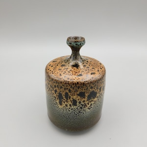 May include: A small, ceramic vase with a speckled brown, orange, and black glaze. The vase has a narrow neck and a wide, rounded base.
