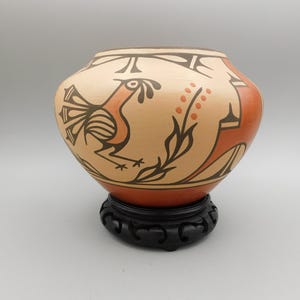 May include: A hand-painted ceramic pot with a rooster design in orange and black. The pot is set on a black wooden stand. The background is a neutral gray.