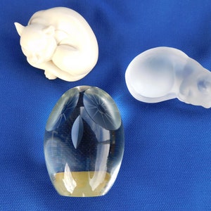 May include: Three decorative figurines on a blue background. A white ceramic cat figurine, a clear glass owl figurine, and a clear glass cat figurine.