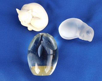 May include: Three decorative figurines on a blue background. A white ceramic cat figurine, a clear glass owl figurine, and a clear glass cat figurine.