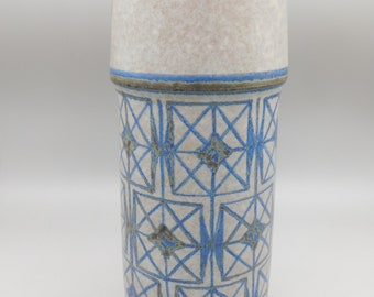 May include: A white ceramic vase with a blue and brown geometric pattern. The vase is cylindrical in shape and has a smooth, glazed finish.