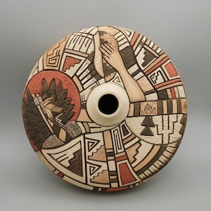 May include: A ceramic vase with a circular design featuring Native American-inspired patterns and imagery. The vase has a cream-colored neck and a central opening. The artwork includes geometric shapes, hands, and a depiction of a bird, in shades of brown, red, and cream.