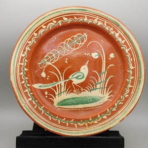 May include: A terracotta-colored ceramic plate with a hand-painted bird and plant design. The bird is white and green, perched on a green base with white and green plants. The rim has a green and white decorative border.