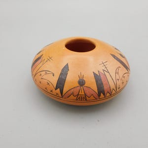 May include: A small, round, orange-colored ceramic pot with a central opening. The pot is decorated with black, red, and brown geometric patterns and shapes. The design includes stylized bird and plant motifs. The pot has a matte finish.