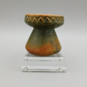 May include: A small, green and orange ceramic vase with a geometric pattern around the top rim. The vase is sitting on a clear acrylic stand.