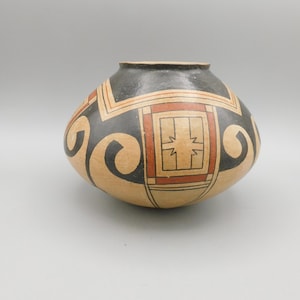 May include: A round, handcrafted pottery vessel with a dark gray rim and a beige body. The vessel features geometric designs in black, red, and beige, including squares and curved lines. The overall design is symmetrical and traditional.