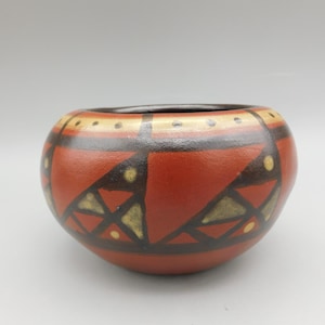 May include: A small, round, terracotta pot with a geometric pattern in black, yellow, and white. The pot is decorated with a repeating pattern of triangles and dots.
