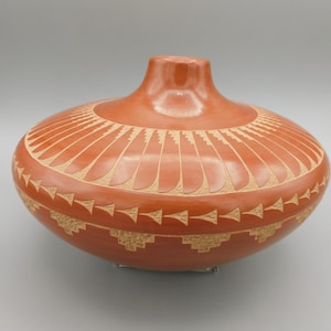 May include: A terracotta-colored ceramic vase with a rounded body and a short neck. The vase features intricate carved designs, including a sunburst pattern near the top and geometric shapes around the base. The vase is approximately 10 inches tall.