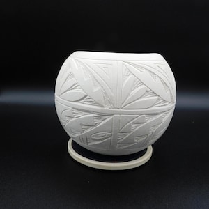 May include: A white, spherical ceramic bowl with a detailed carved geometric pattern. The bowl sits on a small, round, off-white base. The background is a solid black.