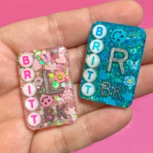 Puede incluir: Dos llaveros rectangulares de resina. Uno es rosa con purpurina, corazones, estrellas y el nombre "BRITT" escrito con cuentas blancas, con las letras "BK" en plata. El otro es azul con purpurina, caras sonrientes y el nombre "BRITT" y las letras "BK" en plata.