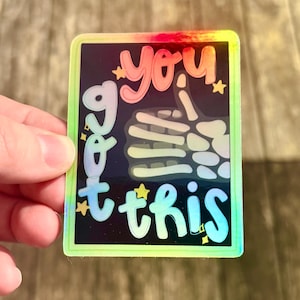 May include: A holographic sticker with the words "you got this" in a colorful, bubble-style font. The design includes a skeleton hand giving a thumbs up and small star accents, all set against a black background.