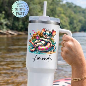 May include: White insulated tumbler with a handle and straw. The tumbler features a cartoon mouse wearing sunglasses and floating on an inner tube. The name "Amanda" is printed on the tumbler. The background shows a river and trees.