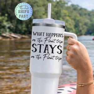 May include: White tumbler with a handle and a straw, featuring the text "WHAT HAPPENS on the Float trip STAYS on the Float trip" in black. The tumbler is held near a river with people floating in the background.