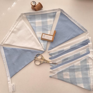 May include: A fabric bunting banner featuring alternating triangular flags in solid light blue, solid white, and a light blue and white gingham check pattern. The flags are attached to a white fabric tape. Includes a wooden spool and scissors.