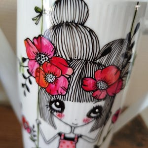 Op de afbeelding: Een witte porseleinen theepot met een zwarte inktillustratie van een figuur met een hoog knotkapsel zittend op een schommel. Het figuur draagt een roze tuinbroek en is omringd door levendige rode klaprozen met zwarte harten en groene stengels.