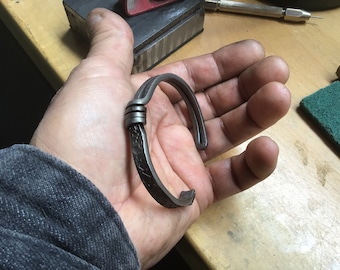 Puede incluir: Un brazalete de metal plateado con un diseño de nudo retorcido. El brazalete está hecho a mano y tiene un acabado martillado.