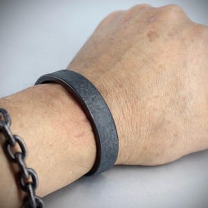 Könnte beinhalten: Ein dunkelgraues, strukturiertes Metallarmband wird am Handgelenk getragen, mit einem passenden Kettenarmband am angrenzenden Handgelenk. Das Armband hat ein flaches, breites Band. Der Hintergrund ist einfarbig hellgrau.