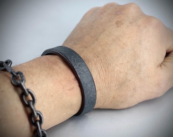 Puede incluir: Una pulsera de metal gris oscuro y texturizado se lleva en la muñeca, con una pulsera de cadena a juego visible en la muñeca adyacente. La pulsera tiene una banda plana y ancha. El fondo es gris claro liso.