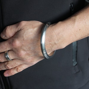 Op de afbeelding: Een zilveren armband met een plat, breed en hoekig ontwerp. De armband wordt gedragen op de pols van een persoon.