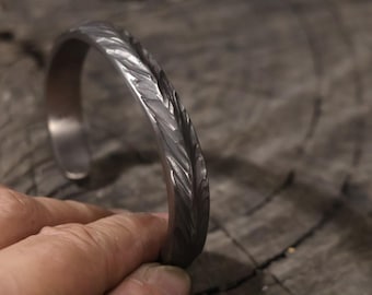 Puede incluir: Un brazalete plateado texturizado. El brazalete de metal tiene un patrón grabado detallado en la parte superior. El brazalete se muestra sobre una superficie de madera.