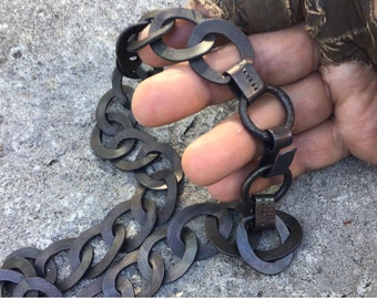Può includere: Collana a catena in metallo grigio scuro con maglie ovali e connettori circolari. La collana è tenuta in alto, mostrando la trama e il design. Il metallo ha un aspetto leggermente usurato.