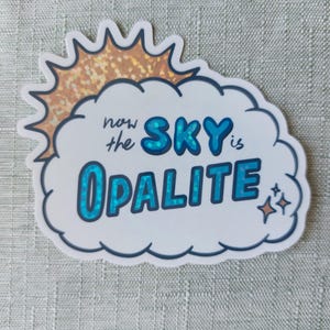 May include: A die-cut sticker featuring a white cloud with a gold holographic sun peeking from behind. The text reads "now the SKY is OPALITE" in blue lettering with a glittery finish, accompanied by small star icons on the right side of the cloud.