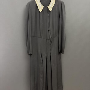 Puede incluir: Un vestido vintage gris oscuro con cuello decorativo. El cuello está adornado con encaje color crema. El vestido tiene mangas largas y una falda plisada. El vestido está colgado de una percha negra.