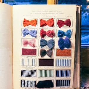 Op de afbeelding: Een opengevouwen vintage boek toont kleurstalen en textielmonsters. De pagina bevat verschillende stoffen strikken in tinten oranje, rood, blauw en paars. Daaronder bevinden zich gestreepte en effen gekleurde stalen. De tekst op de pagina is in het Frans.