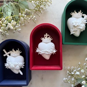 May include: Three heart-shaped religious sculptures in arched wooden frames. The frames are painted in blue, red, and green. The white hearts feature flames, thorns, and floral details. Decorative sprigs of white flowers are arranged around the sculptures.