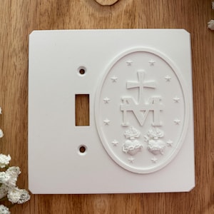 May include: A white light switch cover with an oval emblem featuring a cross, the letters "M" and "I", and two hearts. The cover has a rectangular opening for the switch and two smaller holes.