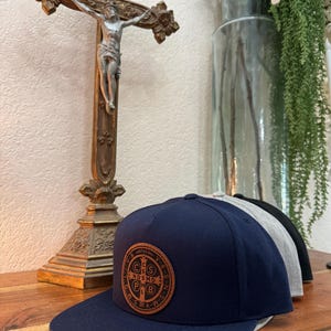 May include: A navy blue baseball cap with a brown leather patch featuring a Saint Benedict medal. The medal has the inscription "CSSMLNDSMDPLB" and "VRSNSMVSMQLIVB", surrounded by a circle of small crosses.