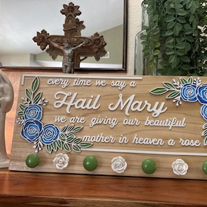May include: Wooden sign with the text "Hail Mary" and floral accents. The sign features blue and white roses, green leaves, and green and white knobs. A statue of Mary and a crucifix are in the background.