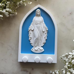May include: White wall hanging featuring a sculpted figure of a woman with outstretched arms, set against a blue backdrop. The figure is framed by a white arch and includes three white knobs below. Decorative religious item.