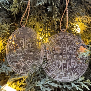 May include: Two clear, circular Christmas ornaments with gold-colored string hangers. One ornament features a cross, hearts, stars, and the letters "M" and "V". The other ornament has a circular design with the letters "PAX" and "C S S M L N D S M D P B".