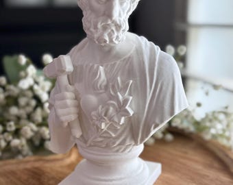 May include: A white bust sculpture of a man with a beard, holding a hammer. The sculpture features a heart and flower design on the chest. The bust is on a square base and set on a wooden tray.