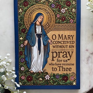 May include: A wooden plaque with a blue frame features a carved image of the Virgin Mary in blue and white robes, surrounded by floral designs. The text "O Mary conceived without sin pray for us" is displayed.