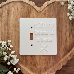 May include: A white light switch cover with the text "St Michael Defend us in Battle" and crossed swords. The cover has a rectangular shape with a switch opening and two screw holes. The cover is on a wooden heart-shaped tray.
