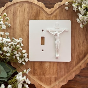 May include: A white light switch cover featuring a 3D depiction of Jesus on the cross. The switch plate has a rectangular shape with a single switch opening and two screw holes. The background is a wooden heart-shaped tray with white flowers.