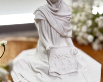 May include: A white statue of a woman wearing a draped head covering and gown, holding a cloth with a detailed face carving. The statue is on a white, geometric base, with a blurred floral background.