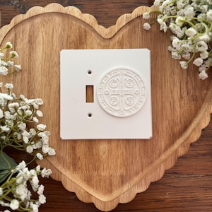 May include: A white light switch cover with a raised, circular emblem featuring religious text and symbols. The cover has a rectangular opening for the switch and two small holes. The switch cover is on a wooden heart-shaped tray.