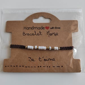 Peut inclure: Un bracelet Morse fait à la main avec un cordon marron et des perles blanches et dorées. Le bracelet est présenté sur une carte marron avec les mots "Handmade with love Bracelet Morse" et "Je t'aime".