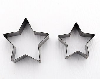 May include: Two metal star-shaped cookie cutters. One is larger than the other. The cutters are made of a shiny, silver-colored metal. They are placed on a white surface.