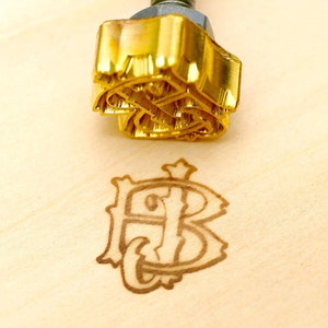 May include: A gold-coloured branding iron with a threaded rod and a nut. The branding iron has a detailed design that has been stamped into the wood. The stamped design is a monogram of the letters "FB".