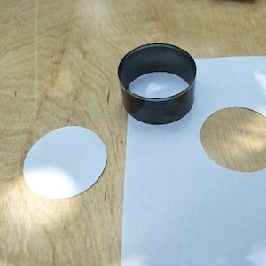 May include: A white paper sheet with two circles cut out. One circle is on the sheet, and the other is on a wooden surface. A metal ring cutter is on the wooden surface.