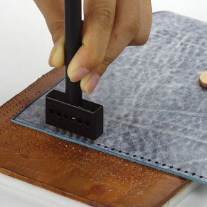May include: A hand using a leather punch tool to create holes in a piece of blue leather. The leather is placed on top of a brown leather surface.