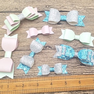 May include: Assortment of 9 glitter bows in various colours, including pink, light blue, silver, and green. The bows are arranged on a wooden surface with a ruler for scale.