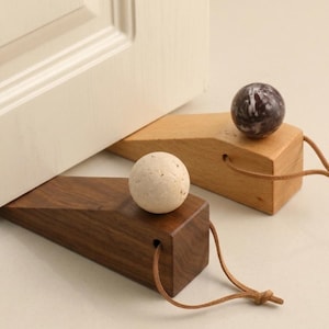 May include: Two wedge-shaped doorstops made of wood, one dark brown and one light tan. Each doorstop features a spherical stone accent on top and a thin leather cord attached to the back. One dark brown doorstop is positioned under a white door.